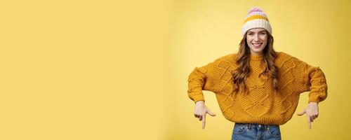 Friendly-looking helpful good-looking 20s college student girl wearing hat warm sweater smiling pleased showing you awesome promotion pointing index fingers down recommend bottom advertisement photo