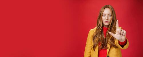Serious-looking redhead woman in yellow coat extending hand to camera with stop and warning gesture prohibiting and forbidding make something having unemotional face as being confident over red wall photo