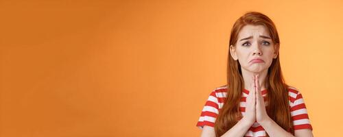 Pity uneasy redhead girl pull sad face, quivering lip upset, begging mercy, apologizing, plead help, press palms pray gesture, frowning hopeless, asking and begging favour, stand orange background photo
