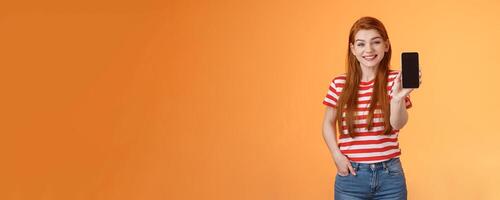 Cheerful good-looking 20s redhead woman introduce social media feature product hold smartphone, show camera cellphone display, promote device application, smiling broadly, orange background photo