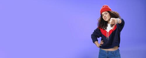 Nah dislike. Portrait of annoyed and unimpressed arrogant cool girl with curly hair tilting head with distain and scorn showing thumbs down in disappointment grimacing, posing over blue wall photo