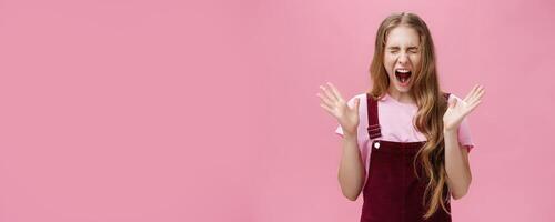 Young girl scared to death seeing spider in room yelling out loud closing eyes and opening mouth gesturing with raised hands near chest feeling afraid and insecure posing against pink background photo