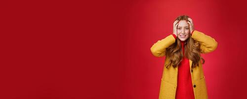 Portrait of redhead woman in panic holding hands pressed to head clenching teeth and frowning puzzled and troubled, standing anxious and concerned freaking out over red background photo
