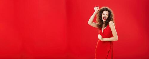 Rich woman forgetting all troubled on dance floor. Charming elegant young european female model in stylish evening dress moving to rhythm of music rejoicing raising hand, singing posing over red wall photo