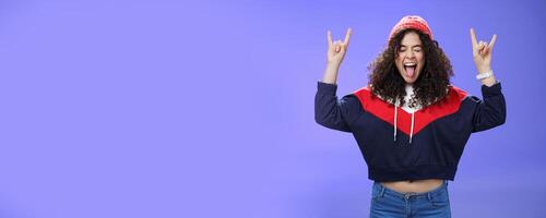 Waist-up shot of rebellious and carefree woman having fun feeling excited and awesome at party close eyes sticking out tongue and showing rock-n-roll signs, wearing beanie and warm clothes photo
