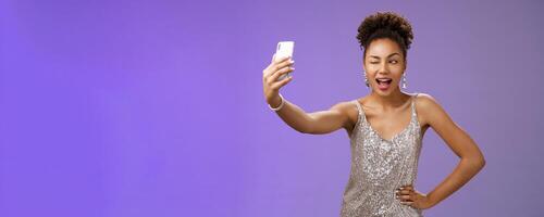 Stylish sassy african american b-day girl taking selfie new elegant silver shiny dress extend arm holding smartphone posing winking display screen amused having fun smiling broadly, blue background photo
