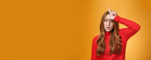 Arrogant and self-satisfied redhead woman humiliating person showing loser sign on forehead mocking and disdain rival standing confident and snobbish over orange background photo