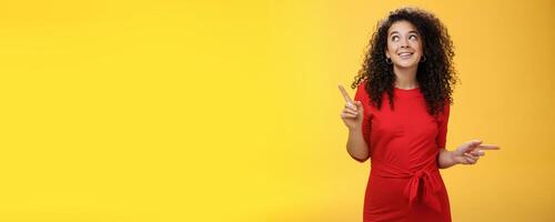 Dreamy charming woman making choice as checking out two awesome products pointing sideways looking intrigued and curious at upper left corner, smiling dreamy and delighted over yellow wall photo