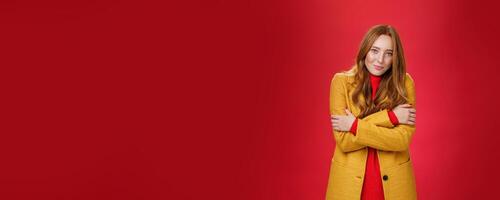 Romantic good-looking tender and cute ginger girlfriend with freckles hugging herself and looking with sensual gentle smile at camera getting cold or chilly waiting outside over red background photo