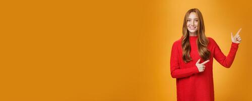 Friendly-looking joyfuly and energized redhead female in red sweater pointing at upper right corner promoting advertisement with broad delighted and tender smile posing against orange background photo