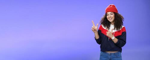 Portrait of friendly good-looking woman with curly hair in red warm beanie pointing at upper left corner and smiling delighted as promoting cool copy space against blue background photo