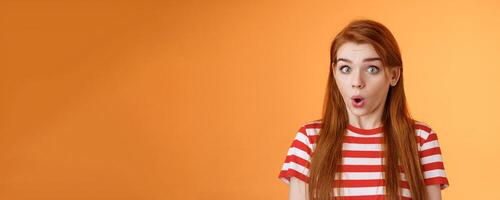 Impressed surprised redhead girl pull astonished face, open mouth gasping amazed, say wow stare camera fascinated, speechless hear awesome promo, stand orange background excited, hear cool news photo