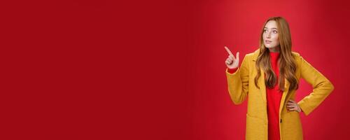 Wondered curious good-looking redhead woman in coat stopping on way looking and pointing at upper left corner curious and interested, observing surprising promotion over red background photo