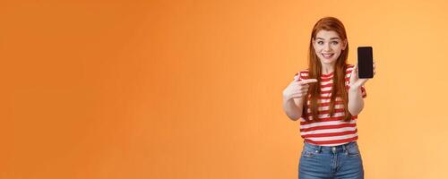 Impressed excited happy redhead woman smiling broadly, hold smartphone, pointing cellphone screen, thrilled amazed introduce fascinating application, stand orange background rejoice photo