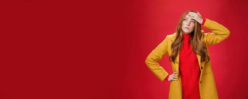 Girl having feaver touching forehead with palm looking displeased and bothered left as thinking she got cold standing in yellow coat, realising she sick or ill over red background, feeling tired photo