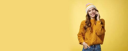 Charming stylish european woman wearing hat sweater tilting head flirty smiling talking smartphone calling boyfriend consulting friend via cellphone, standing happily casual pose yellow background photo