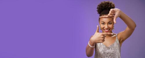 Creative good-looking african woman in silver glittering dress searching inspiration make finger frame look camera pleased smiling satisfied found perfect photo angle, standing blue background