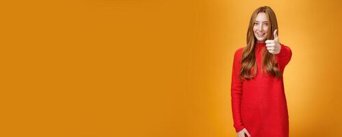 Nice job good. Portrait of delighted and energized confident ginger woman in red sweater pulling thumb up at camera and smiling, liking idea, approving and accepting it over orange background photo