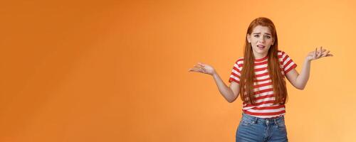 Careless arrogant redhead rich girl say so what, not giving care what people thing, look dismay ignorant shrugging hands spread sideways, uninterested, give no concern, stand orange background photo