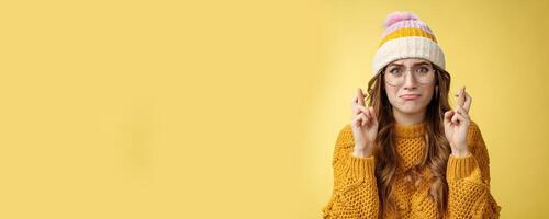 Nervous insecure unconfident cute nerd girl university student wearing glasses sulking cry, whining worried cross fingers good luck praying dream come true standing intense yellow background photo