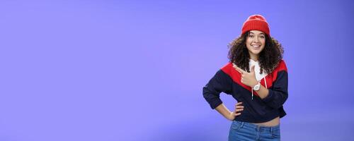 Studio shot of happy charming stylish city girl in beanie and warm sweatshirt ready rock winter up pointing at upper left corner and smiling delighted showing cool place over blue wall photo