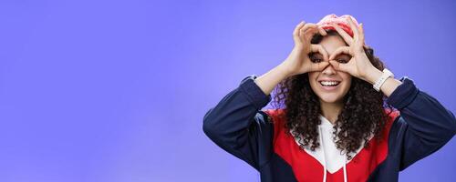 Girl see us as imitating peek with binocular making circles over eyes and looking through it joyful and excited, having fun fooling around posing happy and cute over blue background in outdoor outfit photo