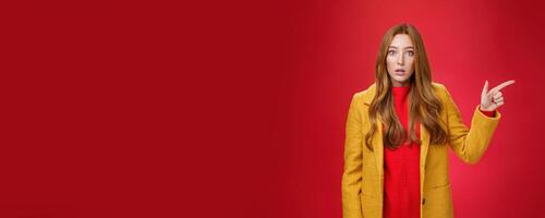 Impressed and questioned amazed redhead woman remain speechless from amazement pointing right open mouth in stupor, asking question about confusing shocking promotion over red wall photo