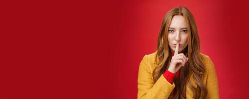 Shh keep out secret safe. Portrait of sensual attractive and daring redhead female in yellow coat showing shush gesture with index finger on lips, squinting looking determined and sassy over red wall photo