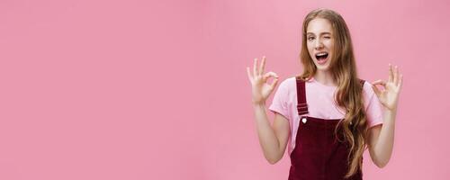 Waist-up shot of assertive good-looking confident teenage girl having everything under control winking delighted and self-assured showing okay gesture confirming job done well over pink background photo