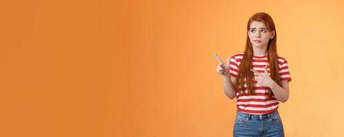 Complicated troubled cute young redhead girl puzzled facing tough difficult decision, look uneasy frowning perplexed hesitating, stare pointing upper left corner disappointed, orange background photo