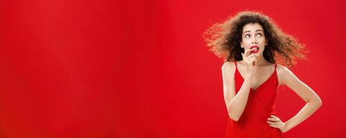 Waist-up shot of concerned and worried insecure cute female with curly hairstyle in red evening dress biting finger looking troubled or questioned at upper right corner with hand on waist photo