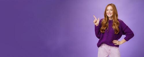 Hey click here. Portrait of friendly-looking charming and cute redhead female in purple sweater pointing at upper left corner with index finger and smiling, showing copy space, recommending, offering photo