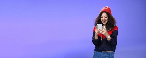 Joyful and sociable stylish caucasian woman with curls in warm beanie having fun laughing and chuckling as using smartphone typing funny message looking at mobile phone screen happily over blue wall photo