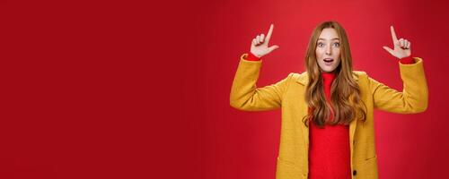 Surprised redhead girl in yellow coat open mouth impressed and thrilled as raising hands and pointing up telling about cool promotion with excited expression against red background photo