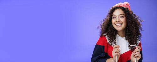 Close-up shot of carefree attractive feminine girl with curly hair in beanie playing with sweatshirt as posing over blue background, smiling at camera having fun and positive attitude photo