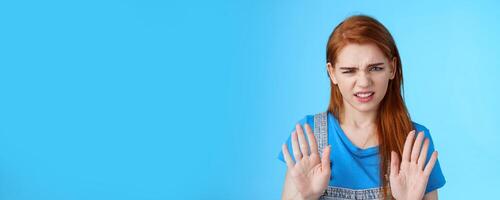 Close-up uninterested displeased ignorant picky redhead girlfriend, frowning make disgusted grimace raise hands block stop gesture, refuse, waving arms no rejection sign, stand blue background photo