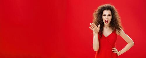 Girl chowing her temper to employees being fed up with unappropriate behaviour complaining standing pissed and dissatisfied gesturing with palm and holding hand on hip over red background photo