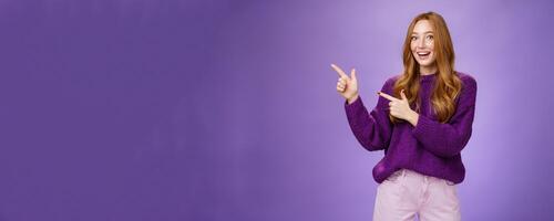 Come in. Portrait of friendly-looking cheerful pretty ginger girl in purple sweater inviting us visit place pointing left with finger pistols and smiling broadly with happy expression over violet wall photo