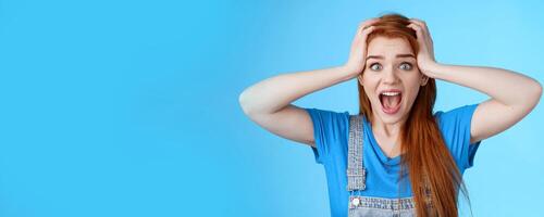 Shocked redhead girl shouting panic, feel pressured deadlines, scared see mess, grab head, scream frightened distressed, stare camera hopeless, anxiously react losing lottery, blue background photo