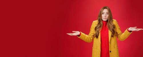 Who new it happen. Innocent and cute redhead unaware girl with freckles in yellow coat shrugging with hands spread sideways, pouting sad and gloomy, being uanware and clueless over red wall photo