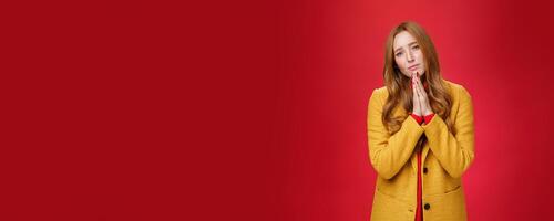 Sorrow and sad attractive redhead girlfriend in yellow coat begging for mercy standing hopeful and upset with hands in pray pouting and frowning, apologizing asking forgiveness over red background photo