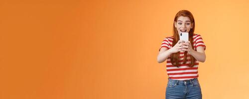 Excited girl recording holding smartphone look through cellphone camera shooting thrilled awesome performance. Cute redhead female student attend cool event use gadget impressed photo