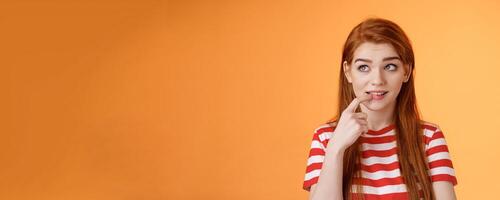 Curious sly cute redhead girlfriend, thinking silly biting finger, look interested upper left corner, have inriguing idea, ponder choice, deciding order online, stand thoughtful orange background photo