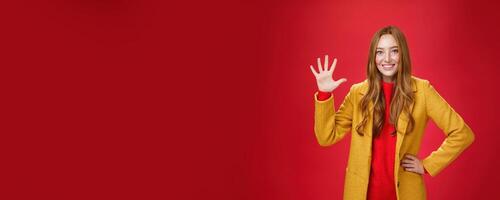Girl giving five stars to awesome internet store she bought outfit for autumn weather showing raised palm smiling broadly as standing in stylish warm yellow coat over red background photo