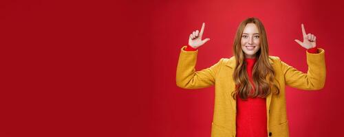 Indoor shot of redhead attractive woman in yellow fall coat raising hands promoting advertisement as pointing up and smiling broadly with satisfied pleasant expression over red background photo