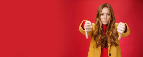 Portrait of insecure and unconfident cute redhead female reacting to uncool thing showing thumbs down with worried upset expression from dislike and disappointment over red background photo