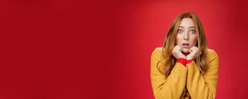 Close-up shot of shocked and scared insecure redhead woman in yellow coat pressing hands to jawline, open mouth and popping eyes at camera from amazement and fear over red background photo