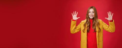 Cute and tender good-looking friendly redhead woman with blue eyes and freckles in stylish yellow coat showing number ten or ordering dozen of things as smiling happily at camera over red background photo