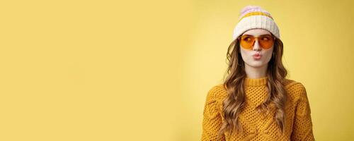 Stylish young glamour thoughtful girl make desicion folding lips silly thinking choosing product look away left wearing sunglasses hat warm sweater enjoying vacation ski resort, yellow background photo