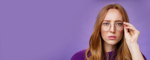 Unsure and serious-looking focused redhead woman frowning and squinting as looking in prescribed glasses at camera, touching frame reading sign, solving puzzle over purple background photo